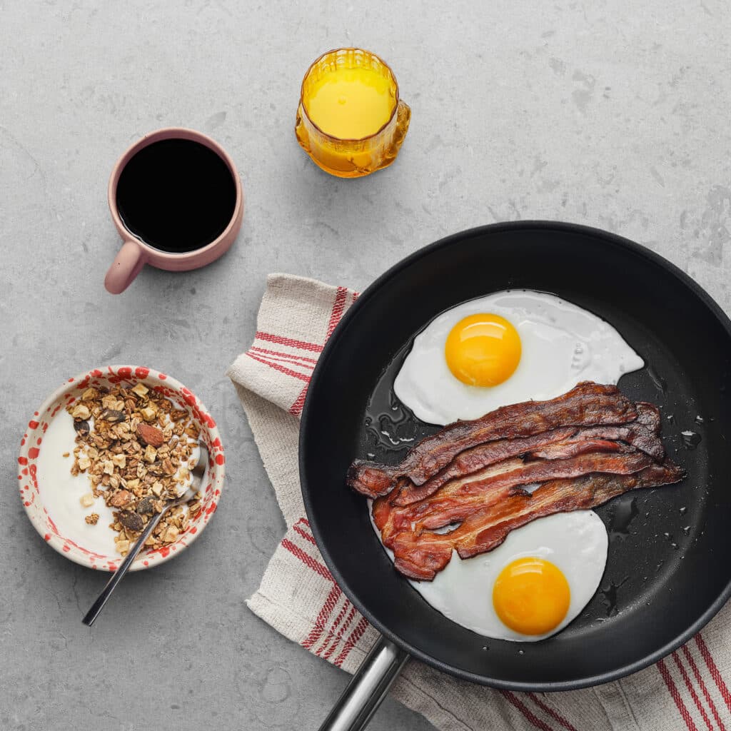 Eggs and bacon in a pan and yoghurt, coffee and juice.